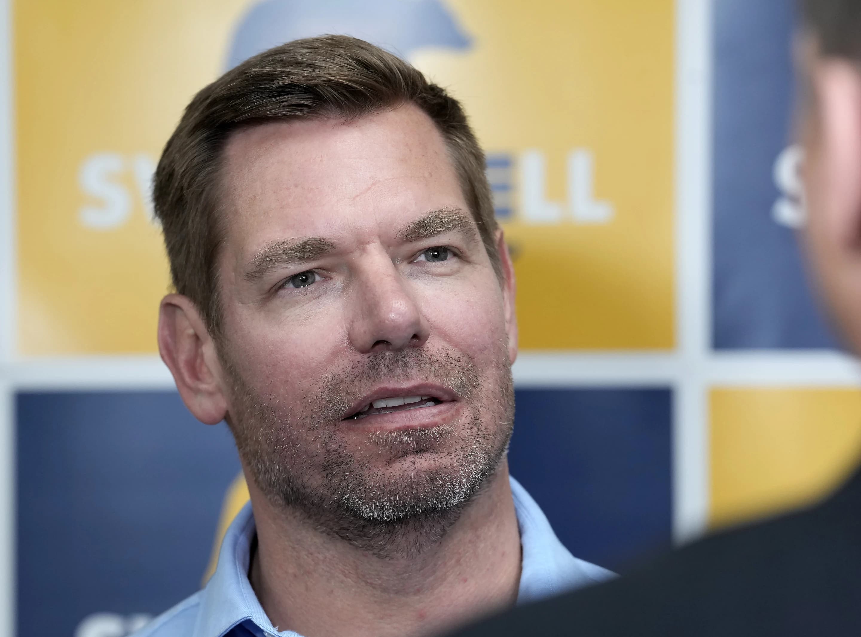 Former Representative Eric Swalwell speaking to reporters in front of a campaign backdrop.