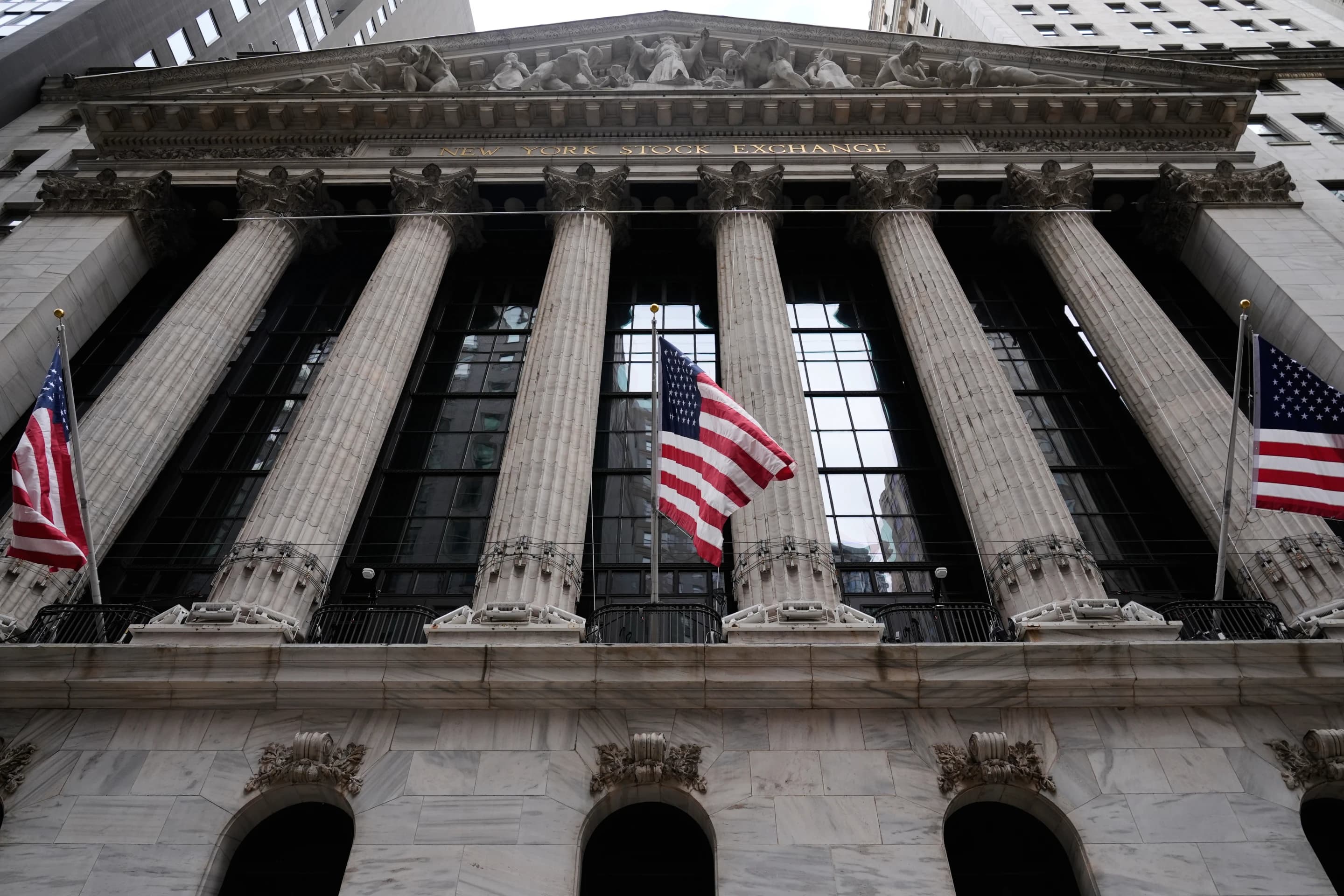 The columned facade of the New York Stock Exchange building with American flags flying in front of it.