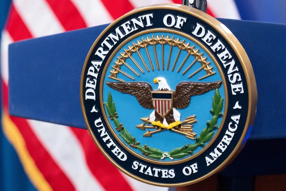The Department of Defense seal mounted on a lectern, with an American flag in the background.