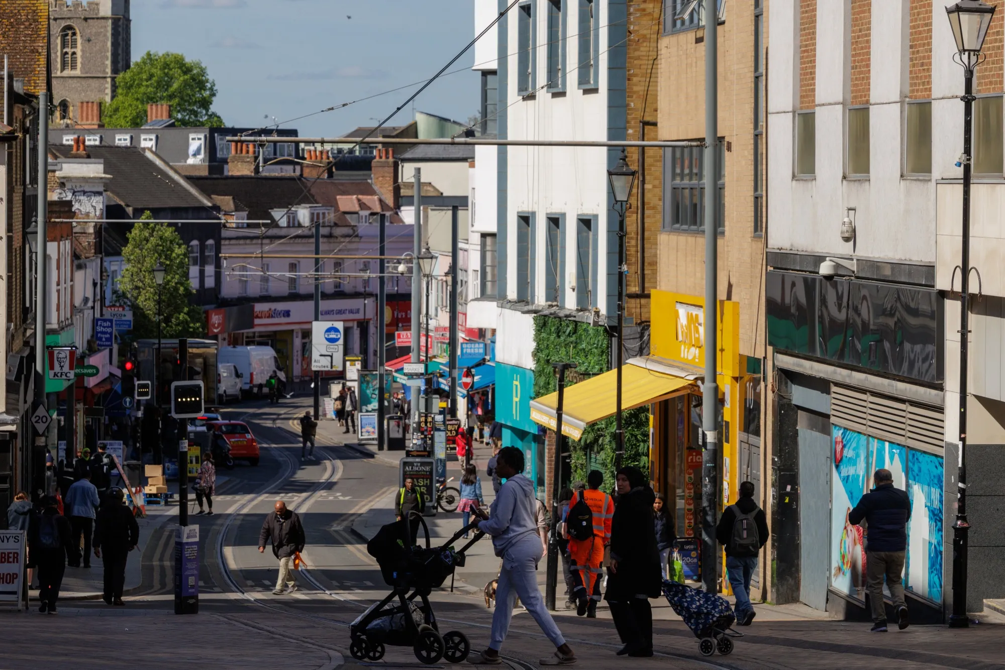 A busy British high street with shops, pedestrians and tram lines in the road.