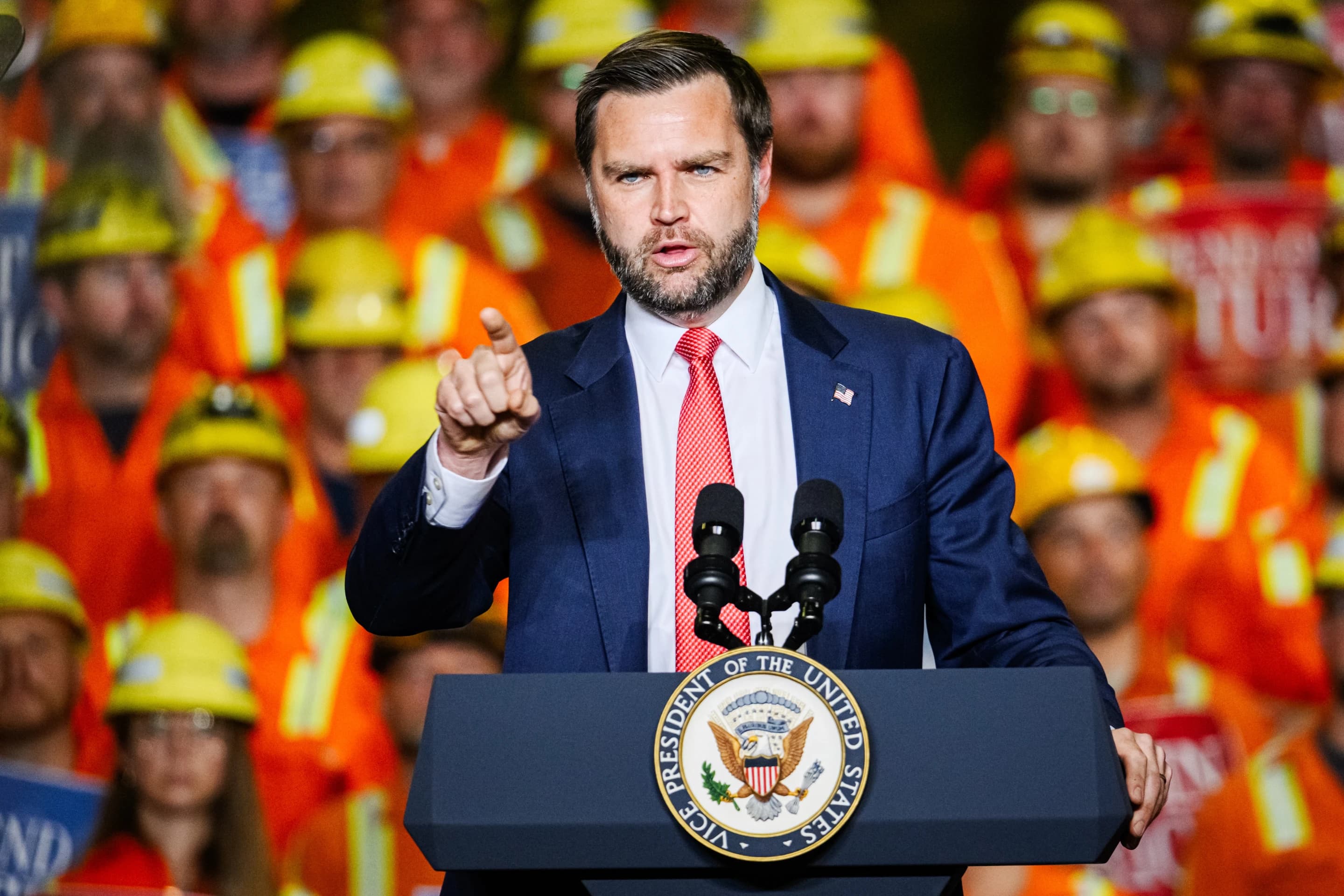 Vice President JD Vance gesturing while speaking at a podium in front of workers wearing orange and yellow safety gear.