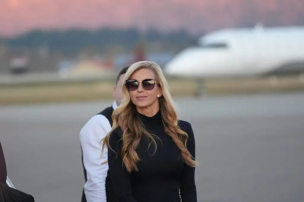Katie Zacharia walking across an airport tarmac wearing sunglasses and a black top, with a plane visible in the background